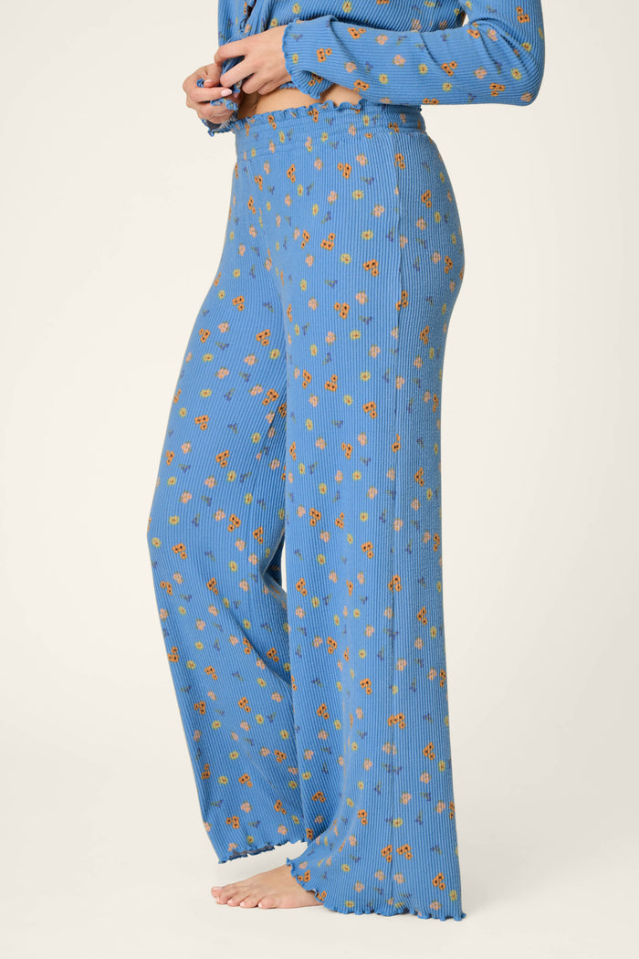A person stands barefoot against a plain light background wearing the P.J. Salvage Tiny Blooms Paris Blue Floral Print Pajama Pants, featuring a blue wide-leg silhouette with a small orange floral pattern.