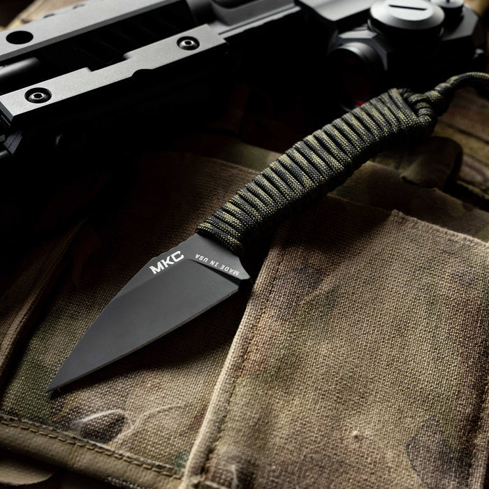 TRAIL GOAT knife with a black blade and dark camo paracord handle, resting on tactical fabric and gear.