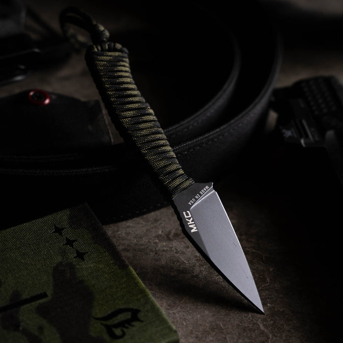 Black and dark camo TRAIL GOAT knife with a paracord handle, displayed on a textured surface with tactical gear.