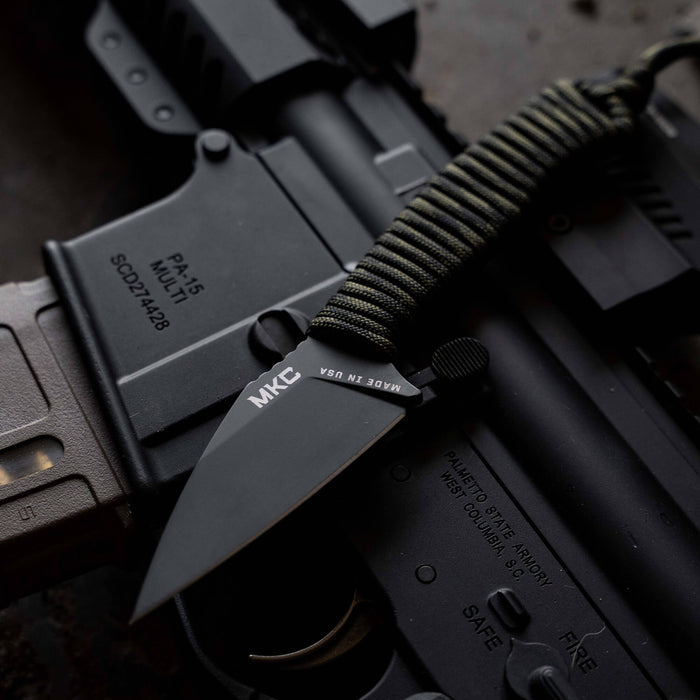 TRAIL GOAT knife with a black blade and dark camo handle, resting on a tactical firearm. Ideal for outdoor use.