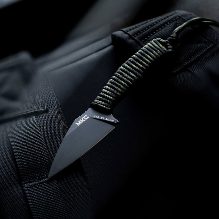 TRAIL GOAT knife with a black blade and dark camo paracord handle, resting on a tactical surface.