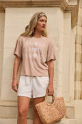 A woman stands against a stone wall wearing a beige T-shirt, white shorts, a headband, and holding a woven straw handbag.