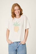 A person with long, wavy, reddish hair stands against a plain background, smiling with both hands in their pockets, wearing blue jeans and the P.J. Salvage Viva la Vida Puff Paint Pineapple T-Shirt.