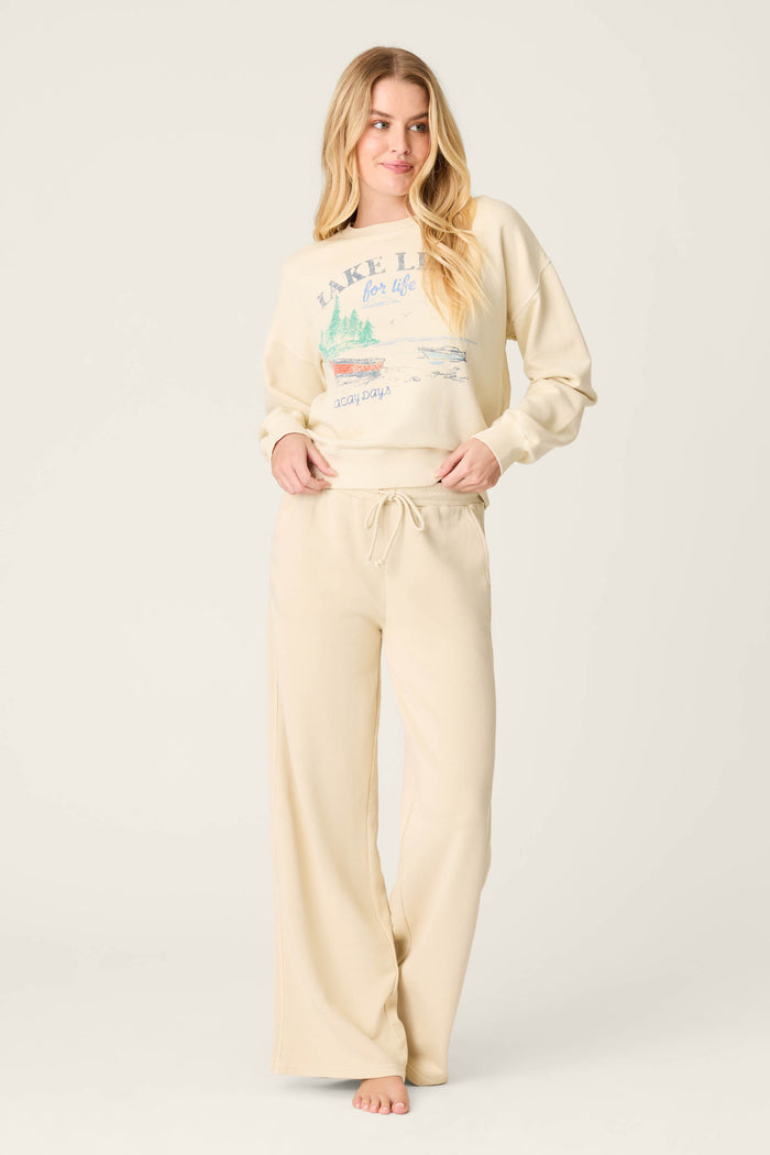 A woman with long blonde hair stands barefoot against a light background, wearing P.J. Salvage's Washed Away Lake Life For Life Open Leg Pants and a matching cream French terry sweatshirt with a graphic in a subtle sun-faded finish.