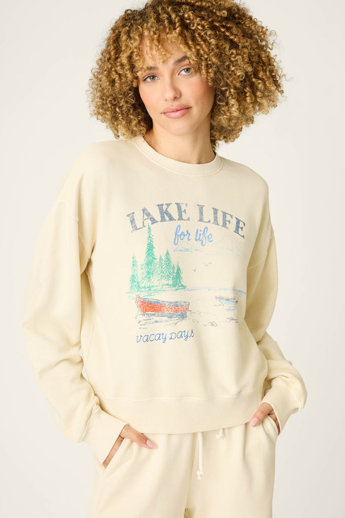 A person with curly hair wears the P.J. Salvage Washed Away Lake Life For Life Pullover Sweater, cream-colored with a canoe, trees, water, and birds graphic and a relaxed fit, paired with matching pants featuring side pockets.