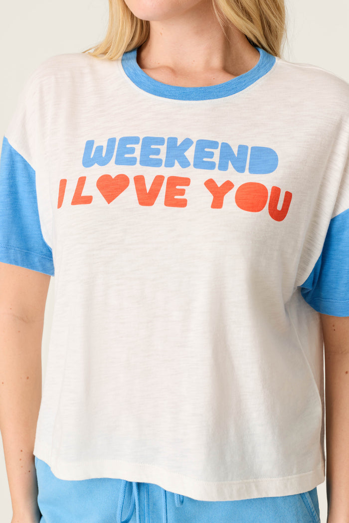 Someone wears the P.J. Salvage Washed Away Weekend I Love You Graphic Tee, a white and blue slub-jersey crewneck featuring "WEEKEND I LOVE YOU" in blue and red letters; their face is partly out of frame, creating a laid-back look.