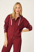 A woman smiles softly while wearing the P.J. Salvage Weekday Waffles Lisette Zip-Up Hoodie in maroon, paired with matching pants featuring white drawstrings and stitched detailing, standing against a plain light background.