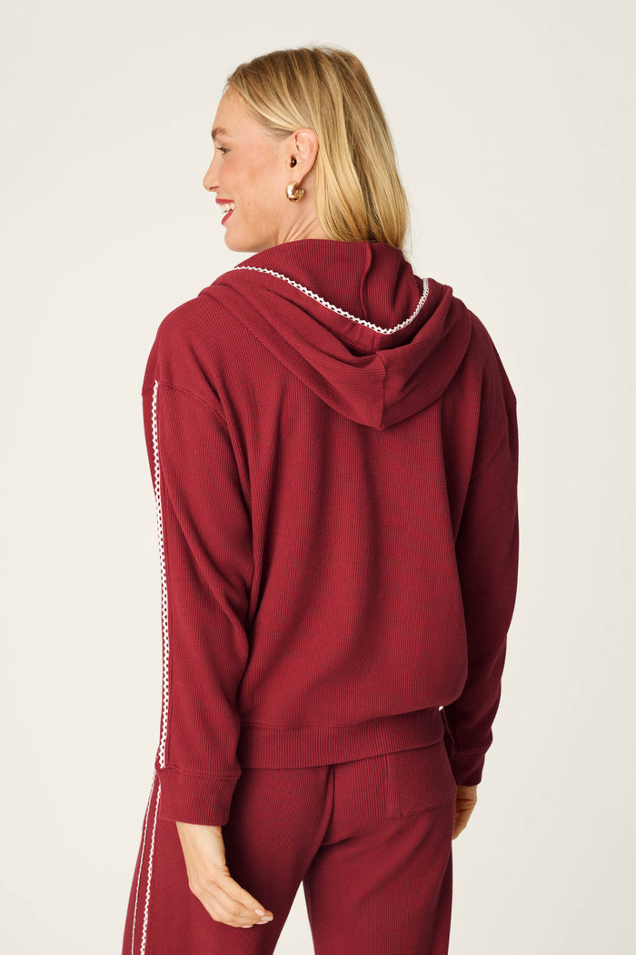 A woman with blonde hair is seen from behind, smiling and showcasing the back of the P.J. Salvage Weekday Waffles Lisette Zip-Up Hoodie in maroon, featuring white stitching along the sleeves, set against a plain background.
