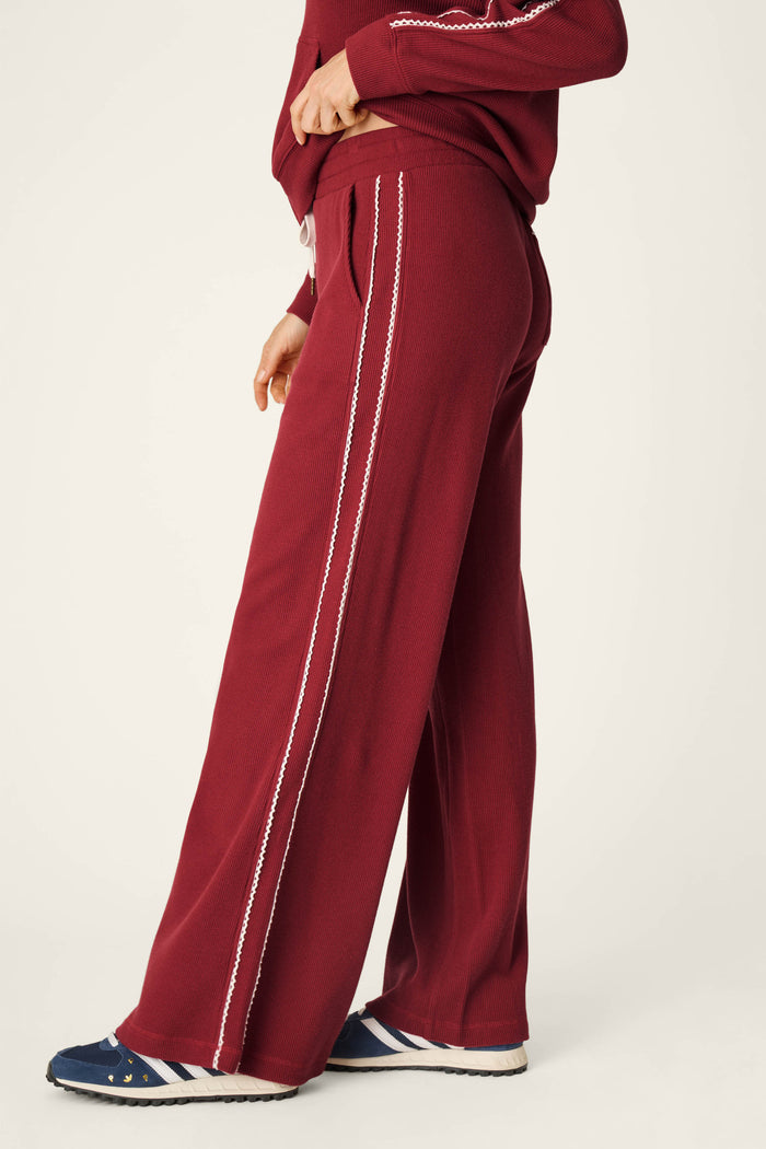Wearing the P.J. Salvage Weekday Waffles Rue Lounge Pants in maroon, paired with a matching top and blue striped sneakers, this person stands sideways against a plain background.