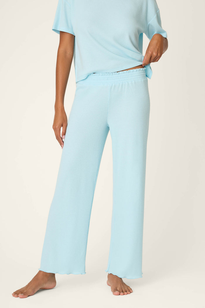 A person stands barefoot against a plain background, holding the waistband of P.J. Salvage’s Weekend Waffles Crystal Blue Pajama Pants in a wide-leg fit, paired with a matching short-sleeve top. Their face is not visible.