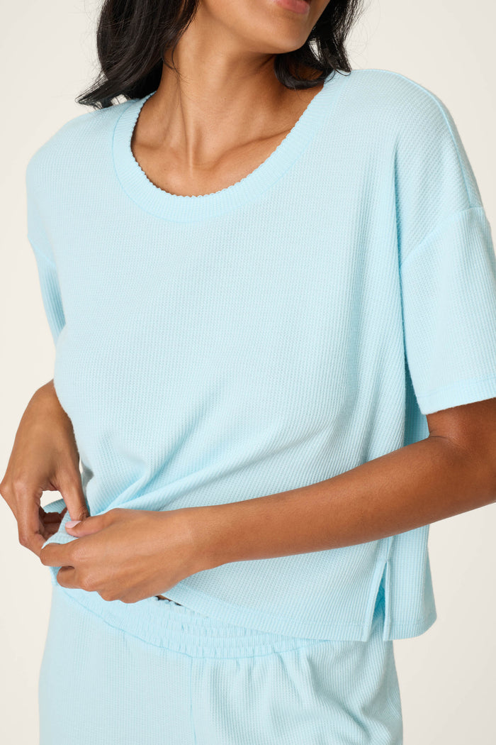 A woman in the P.J. Salvage Weekend Waffles Crystal Blue Long PJ Set tugs at her top. The relaxed set includes a loose-fitting tee and matching elastic waist shorts, perfect for cozy lounging.