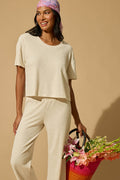A woman in the P.J. Salvage Weekend Waffles Light Oatmeal Long PJ Set and a pink-patterned headscarf smiles while holding a colorful woven bag of flowers against a tan background—ideal for cozy, effortless style.