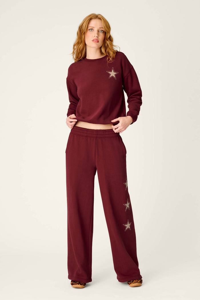 A woman poses in P.J. Salvage’s Western Ways Dark Wine BFF Lounge Pants, featuring star designs, paired with a matching maroon sweatshirt against a plain light background.