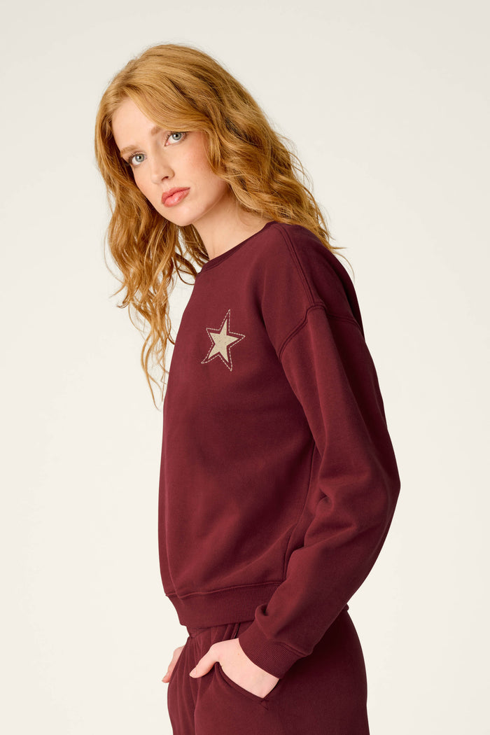 A woman with long wavy red hair stands sideways, looking toward the camera against a plain light background. She is wearing the P.J. Salvage Western Ways Dark Wine Simone Sweatshirt, which features a star patch on the chest, paired with matching pants.