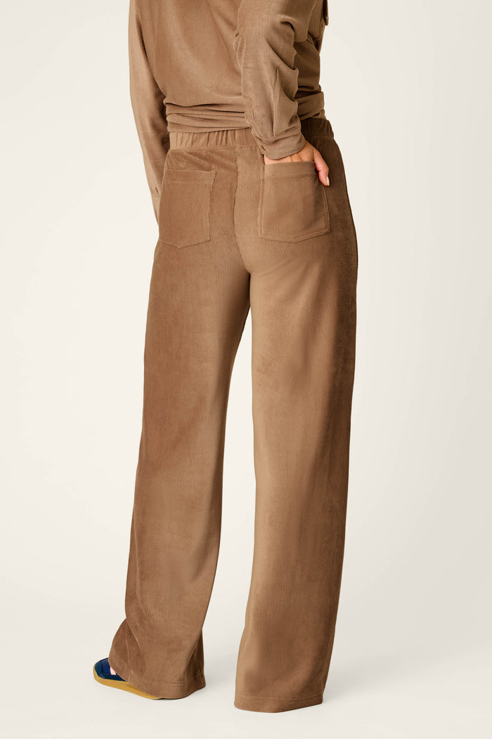 A person is seen from behind against a plain, light background, wearing P.J. Salvage’s Western Ways Easy Itzy Corduroy Knit Lounge Pants in brown with a matching top, standing with one hand tucked into the back pocket.