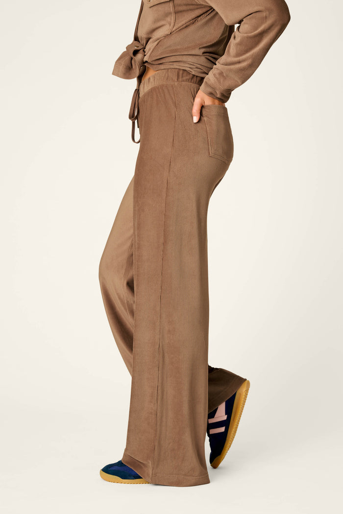 A person is wearing the P.J. Salvage Western Ways Easy Itzy Corduroy Knit Lounge Pants in a loose fit, paired with navy sneakers featuring yellow soles, standing in profile with one hand in their pocket against a plain background.