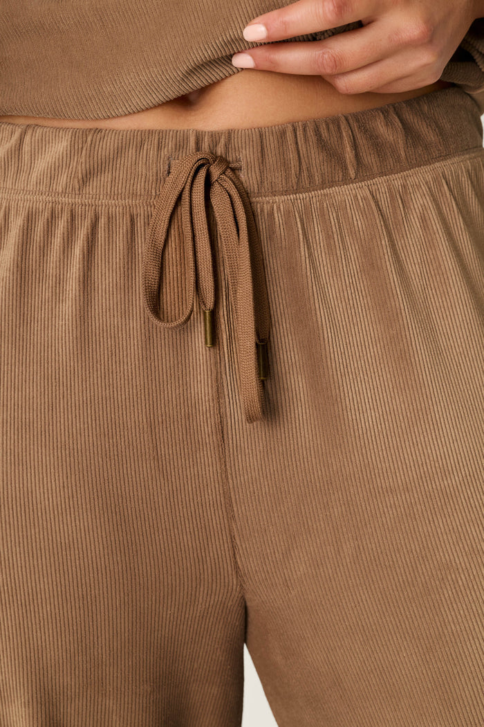 Close-up of a person wearing P.J. Salvage Western Ways Easy Itzy Corduroy Knit Lounge Pants in light brown, featuring a ribbed texture and drawstring waist, with only the hand, waist, and hips visible as they partially lift a matching top.