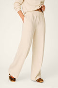 A person wearing the P.J. Salvage Western Ways Good Luck Club Runway Lounge Pants in cream, featuring wide legs and an elastic waistband, paired with a matching top, stands with one hand in their pocket. Leopard print slippers can be seen partially.