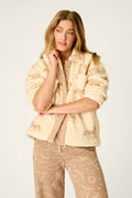 A woman with long, wavy hair wears the P.J. Salvage Western Ways Horse Print Big Teddy Sherpa Jacket—a cream-colored fuzzy layer—over a beige outfit featuring animal and celestial prints, standing against a plain light background.