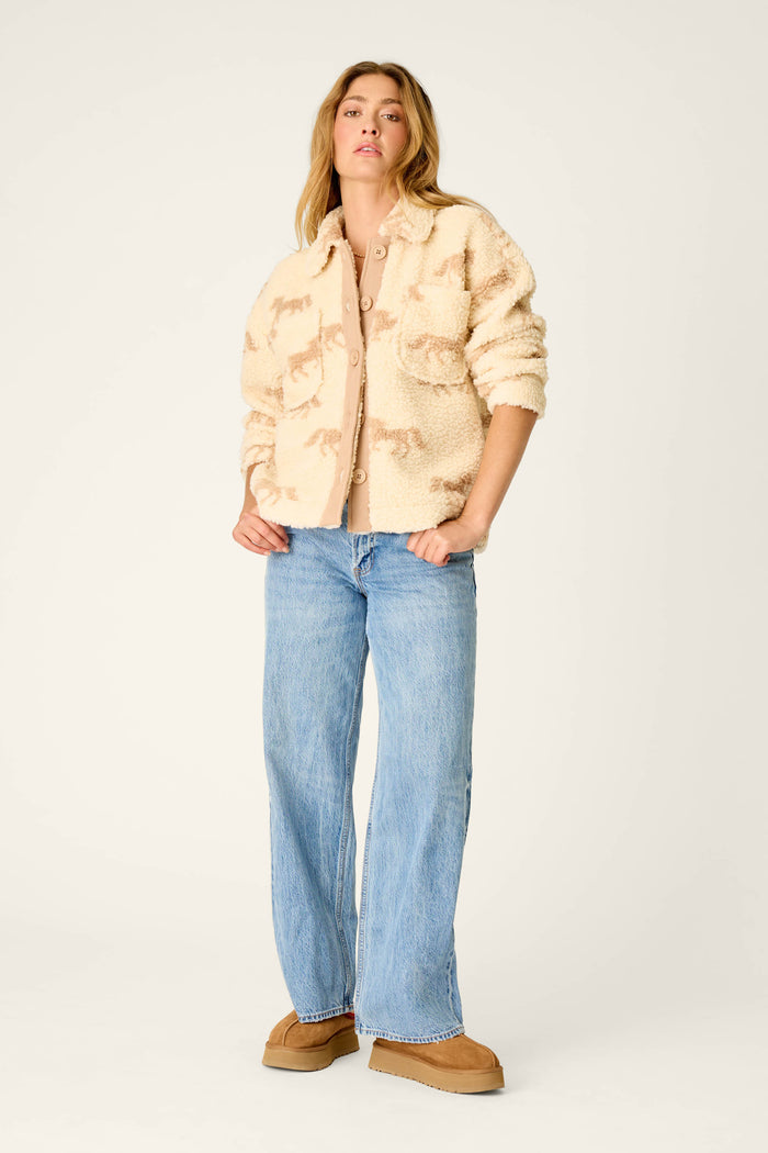A person stands against a plain light background wearing the P.J. Salvage Western Ways Horse Print Big Teddy Sherpa Jacket in beige, styled with a light tan shirt, loose blue jeans, and tan platform shoes.