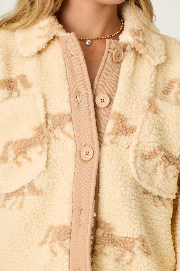 A close-up of a woman wearing the P.J. Salvage Western Ways Horse Print Big Teddy Sherpa Jacket, featuring a cream-colored sherpa fabric with tan horse patterns, large front pockets, and tan buttons. She accessorizes with a delicate gold necklace with a small pendant.