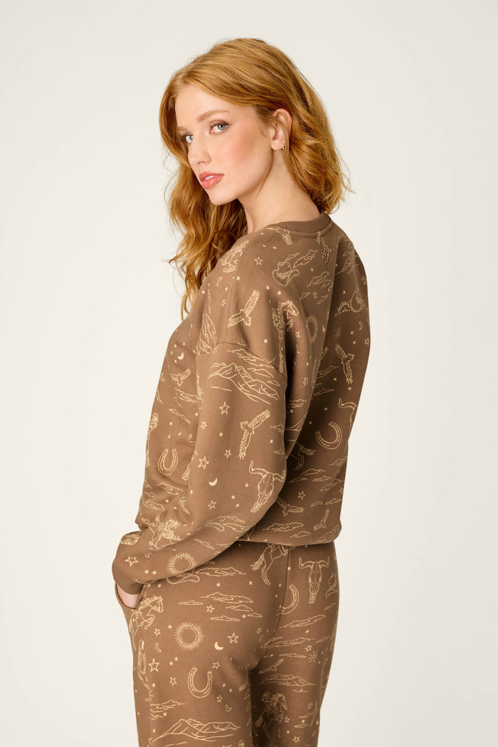 A woman with long red hair stands in profile, looking at the camera while wearing the P.J. Salvage Western Ways Ride ’Em Cowgirl Frankie Sweatshirt—a brown set featuring tan western-themed patterns.