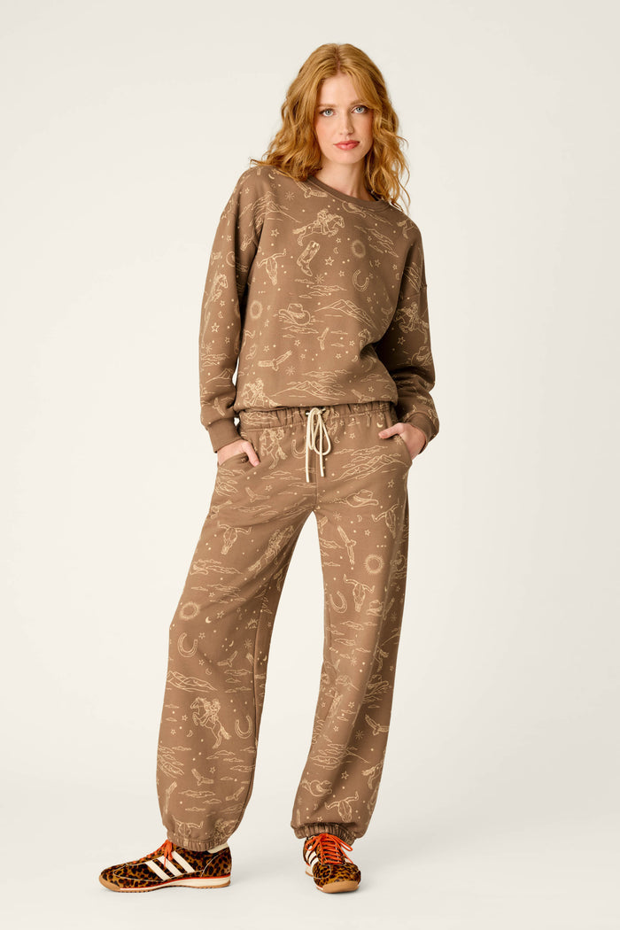 A woman with wavy blonde hair wears the P.J. Salvage Western Ways Ride ’Em Cowgirl Good Hang Joggers—a loose brown sweatsuit with light patterns—paired with brown-and-white Adidas sneakers featuring leopard-print accents, standing against a plain off-white background.