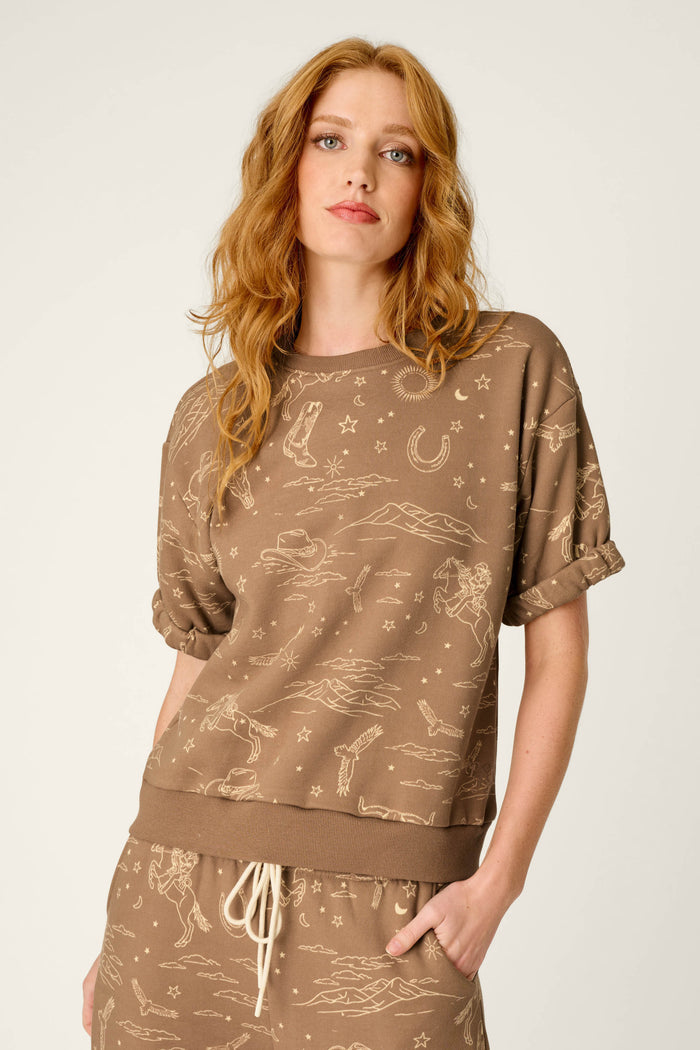 A woman with long red hair wears the P.J. Salvage Western Ways Ride ’Em Cowgirl Roll Back Short Sleeve Sweatshirt and matching pants, featuring beige Western-themed line drawings, while standing against a plain light background.