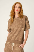 A woman with long red hair wears the P.J. Salvage Western Ways Ride ’Em Cowgirl Roll Back Short Sleeve Sweatshirt and matching pants, featuring beige Western-themed line drawings, while standing against a plain light background.