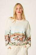 A woman with long blond hair wears the P.J. Salvage Western Ways Untamed Spirit Frankie Sweatshirt, which features a colorful prairie scene with horses and hills. She stands against a plain, light background.