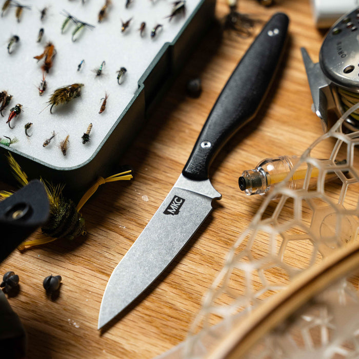 WESTSLOPE knife with a black handle on a wooden surface surrounded by fishing flies and tools.