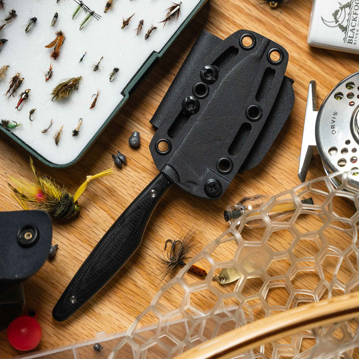 WESTSLOPE knife in black with sheath, surrounded by fishing flies and gear on a wooden surface.