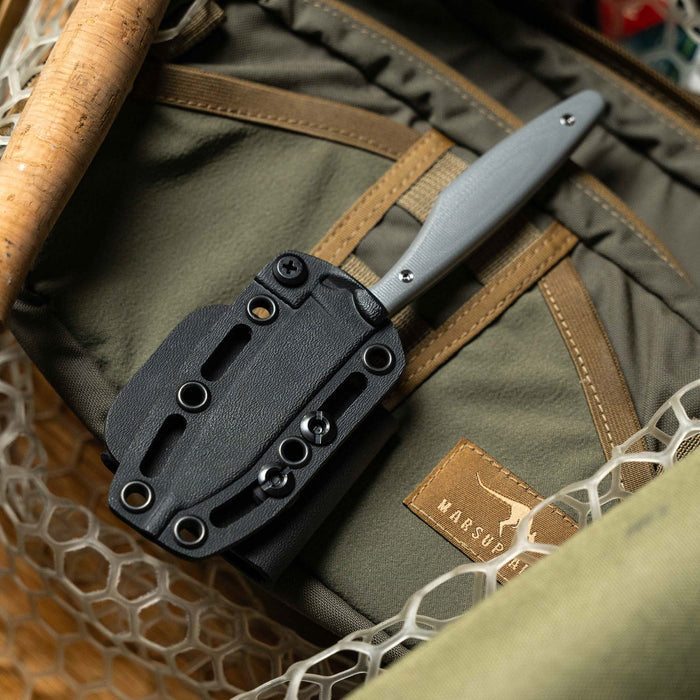 Montana Knife Company's WESTSLOPE knife in grey, secured in a tactical sheath, resting on a backpack.