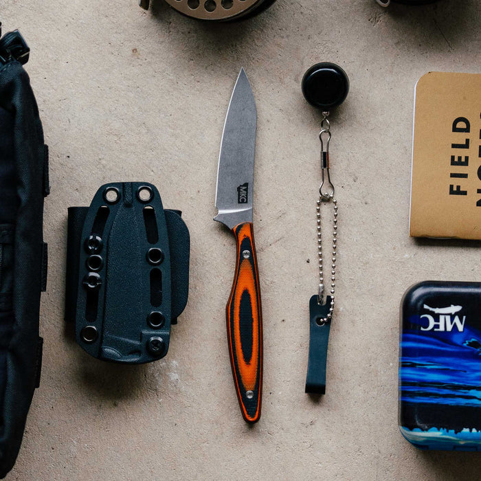 WESTSLOPE knife in orange and black with a fixed blade, sheath, keychain, and notebook on a textured surface.