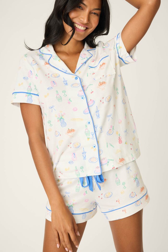 A woman in the P.J. Salvage Whimsy Parisian Picnic Short PJ Set smiles at the camera, wearing a multicolored, short-sleeve button-up top with blue piping and matching shorts with a blue drawstring. Her left arm is raised.