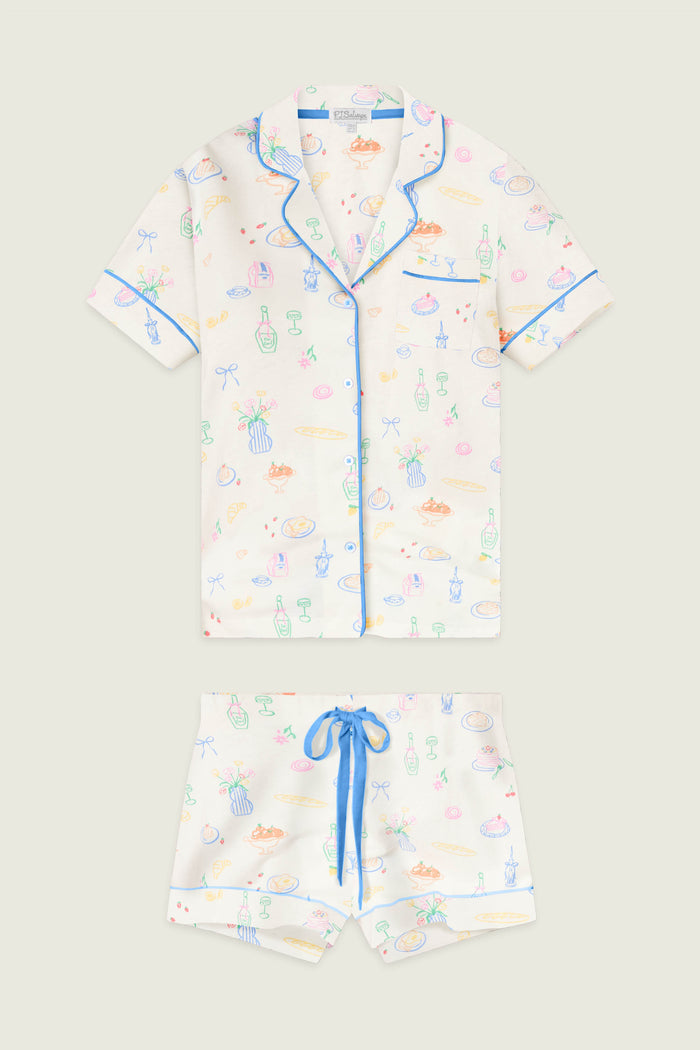 The P.J. Salvage Whimsy Parisian Picnic Short PJ Set features a white short-sleeve button-up top and shorts with blue trim and waist tie, adorned with colorful bottles, pastries, and ribbon prints.
