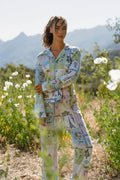 A woman stands in a wildflower field under clear skies, wearing the P.J. Salvage Whimsy Postcards From Paris PJ Set with playful illustrations; mountains rise in the distance.