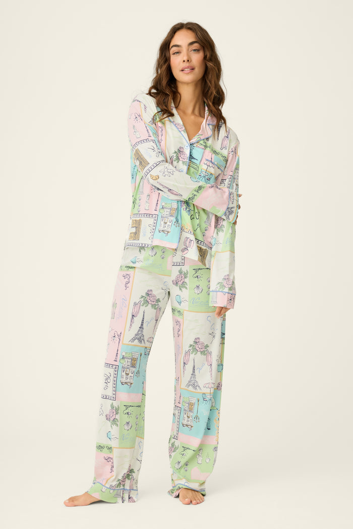 A woman with long brown hair stands barefoot against a light background, wearing the P.J. Salvage Whimsy Postcards From Paris PJ Set—pastel, long-sleeve pajamas featuring a whimsical illustrated print. One arm rests on her torso.