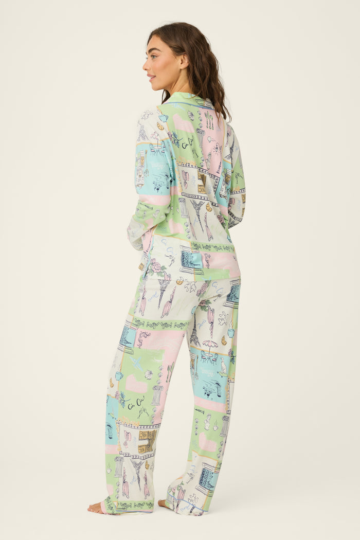 A woman stands barefoot facing away in the Whimsy Postcards From Paris PJ Set by P.J. Salvage, featuring pastel illustrated print, relaxed fit, a long-sleeve button-up top, and loose pants against a plain off-white background.