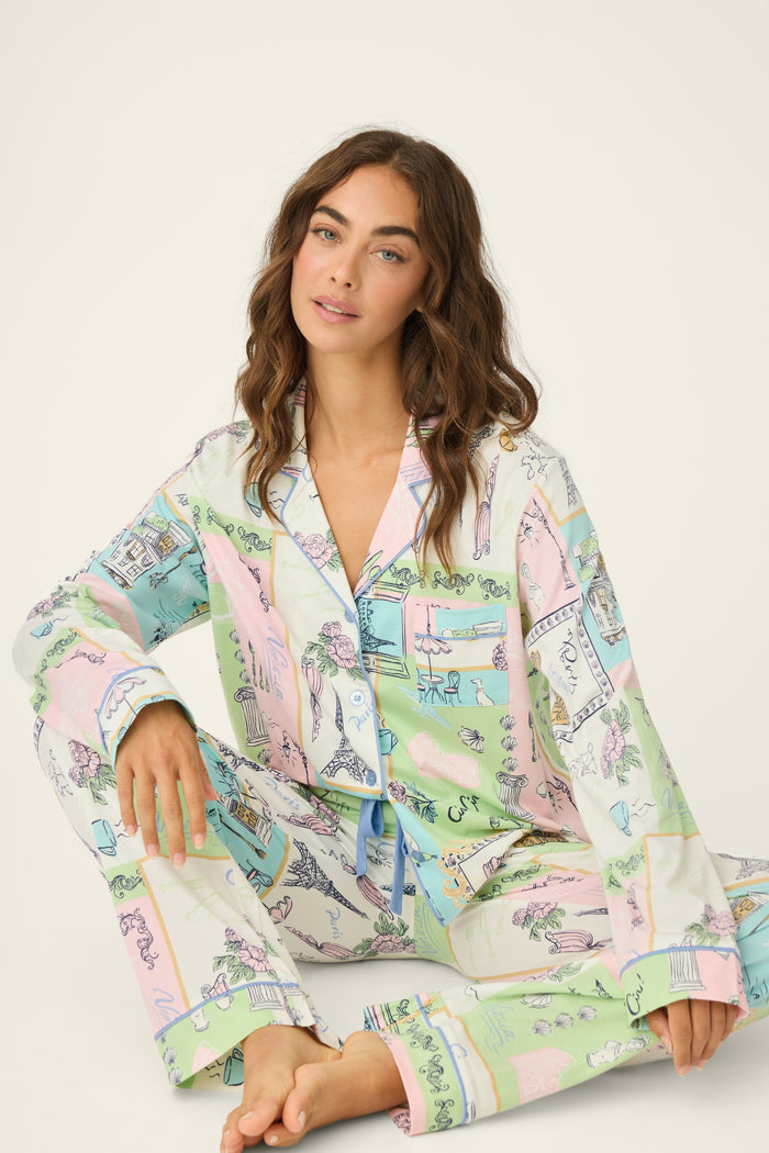 A woman with long brown hair sits on the floor wearing the P.J. Salvage Whimsy Postcards From Paris PJ Set—pastel patterned, button-up shirt and matching pants with charming illustrations in soft tones.