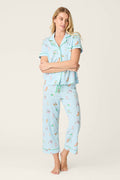 A woman stands barefoot in the P.J. Salvage Whimsy Putter Pup Cropped PJ Set—a light blue pajama featuring a short-sleeve button-up top and long pants with small colorful illustrations—against a plain light background.