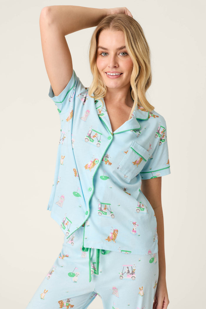 A smiling woman with one arm raised wears the P.J. Salvage Whimsy Putter Pup Cropped PJ Set—light blue pajamas with playful animal and golf cart print, green piping, and buttons—against a plain, neutral background.