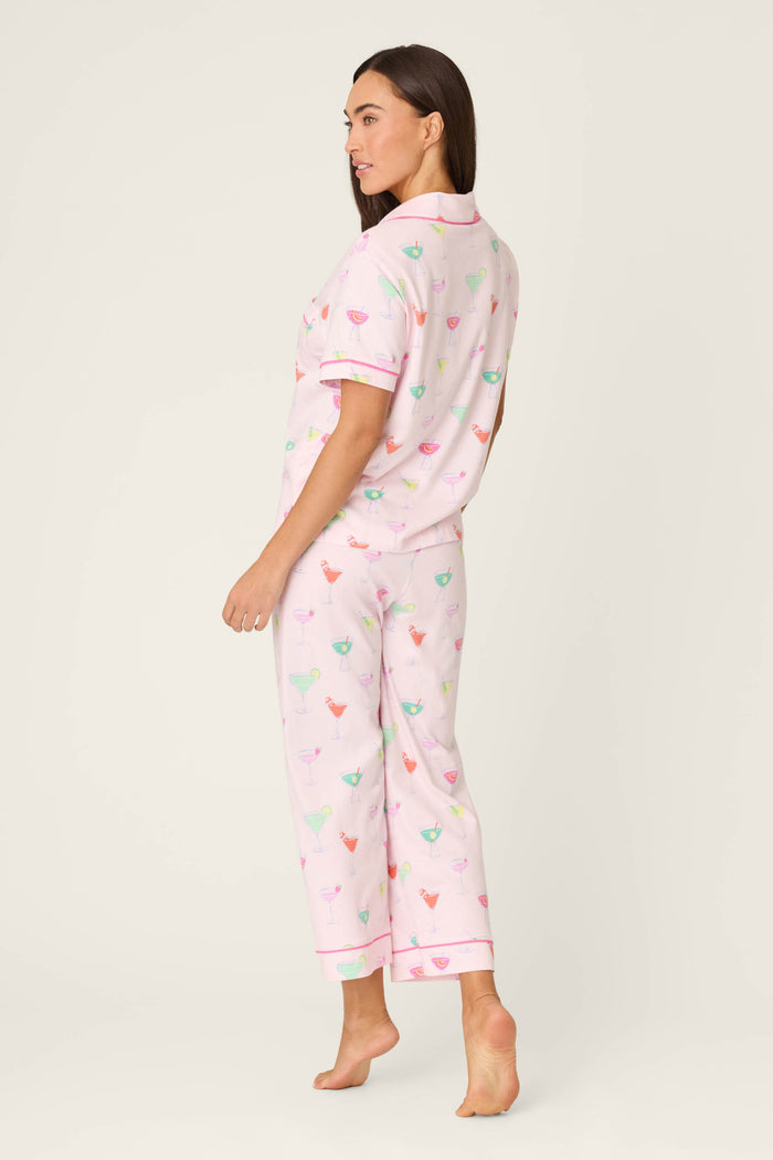 A woman stands barefoot on a plain background, wearing the P.J. Salvage Whimsy Sip Happens Cropped PJ Set featuring a colorful cocktail glass print. She is turned slightly to the side, looking over her shoulder.